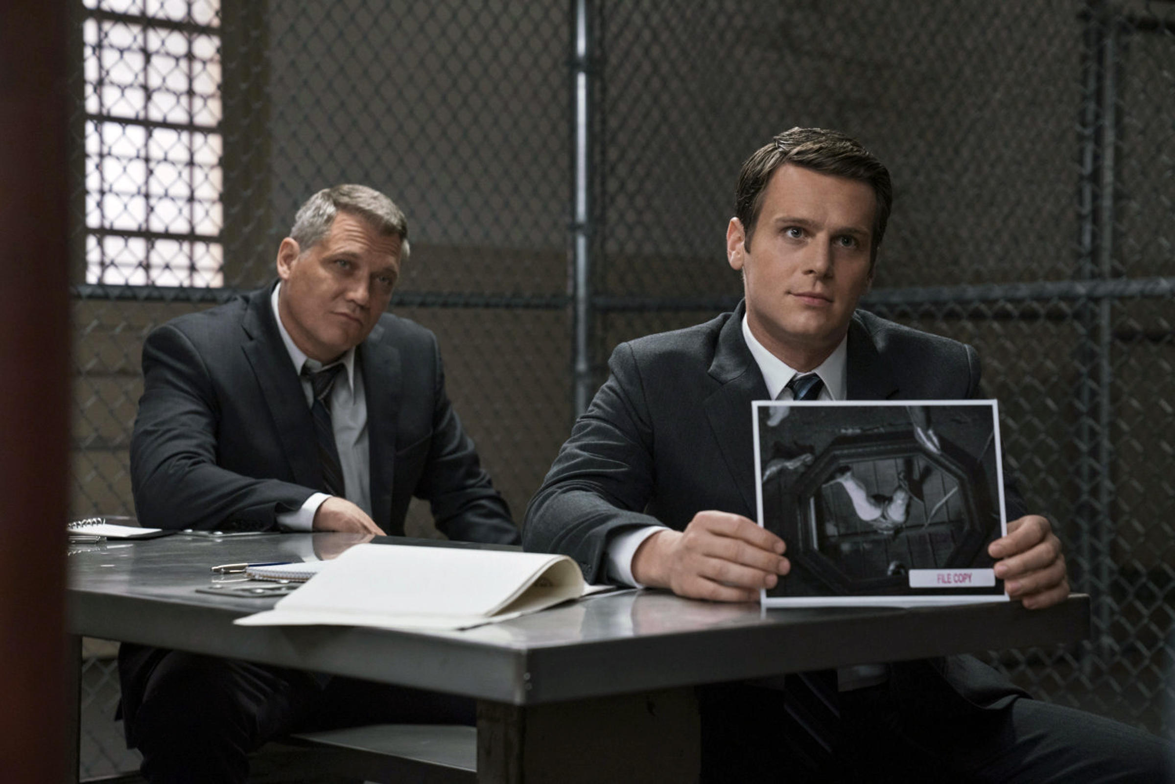 two detectives in suits sit in a jail cell showing pictures in a still from mindhunter