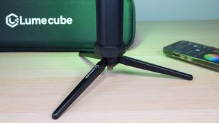 Lume Cube RGB Tube Light Mini on the included stand, emitting a green light, with Lume Cube case in the background and a phone with the Lume Control app