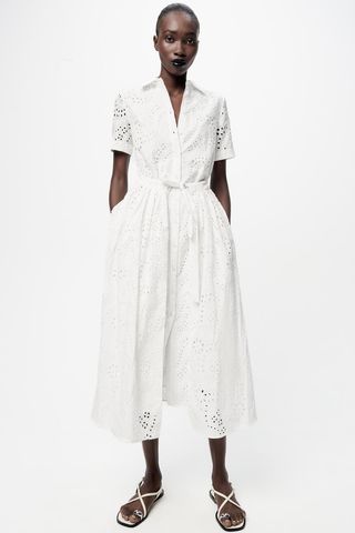 A model wears a shite eyelet shirt dress with white strappy sandals against a white seamless background