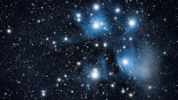 A collection of blue-white stars glow against a field of dimmer stars in the blackness of space. A faint nebulosity can be seen glowing before the brightest stars.