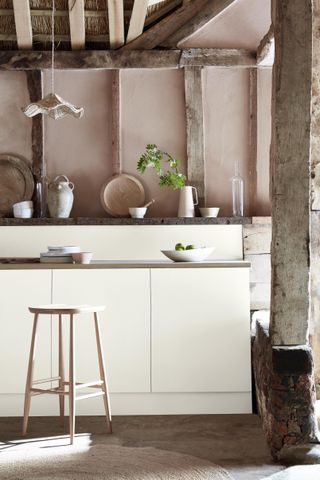 A rustic kitchen with exposed wooden beams, cream units and pink walls