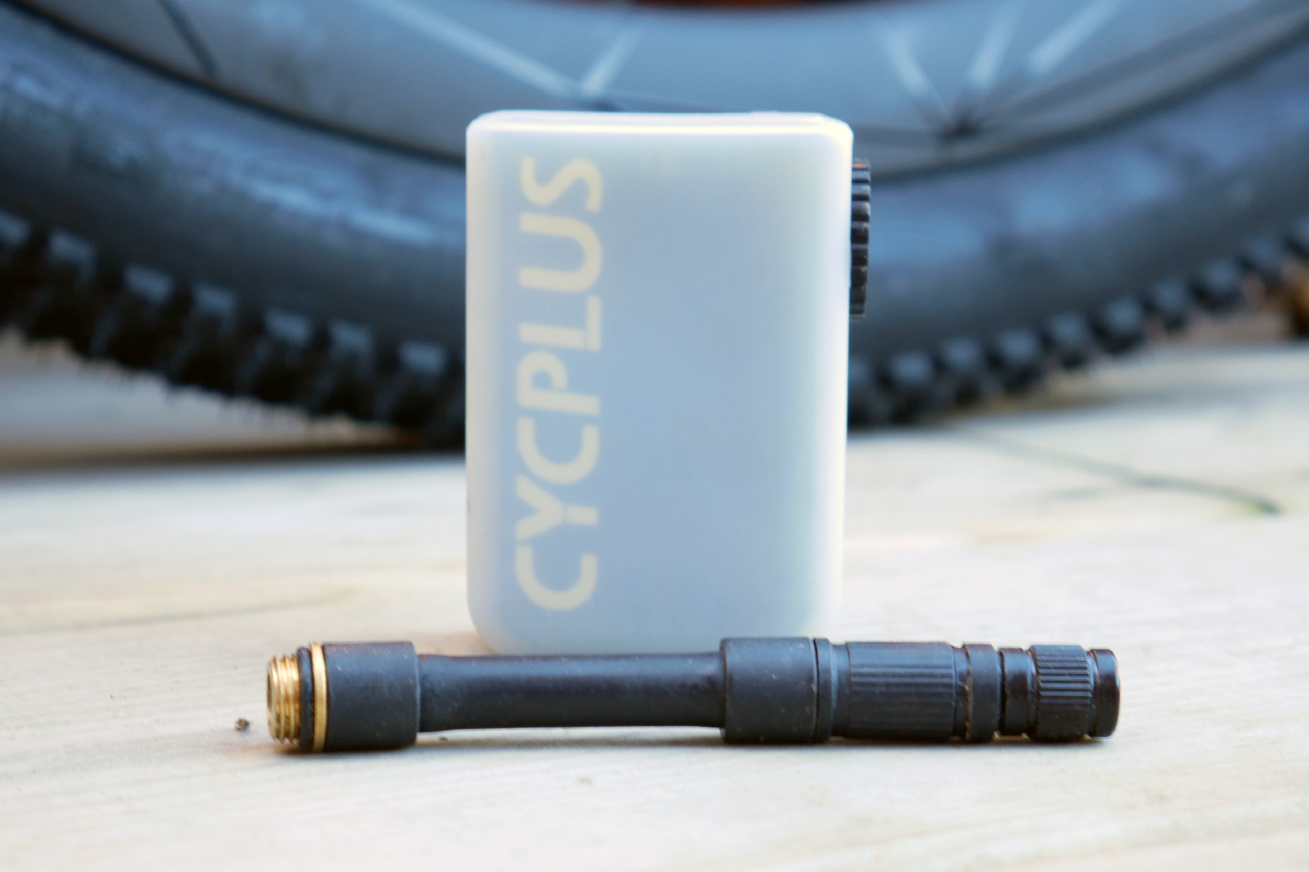 A Cycplus AS2 Ultra electric pump with its valve extension placed on a wooden deck, with a gravel tyre in the background