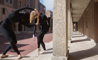 Two woman wearing black dance clothing in a dancing pose in courtyard between some buildings.