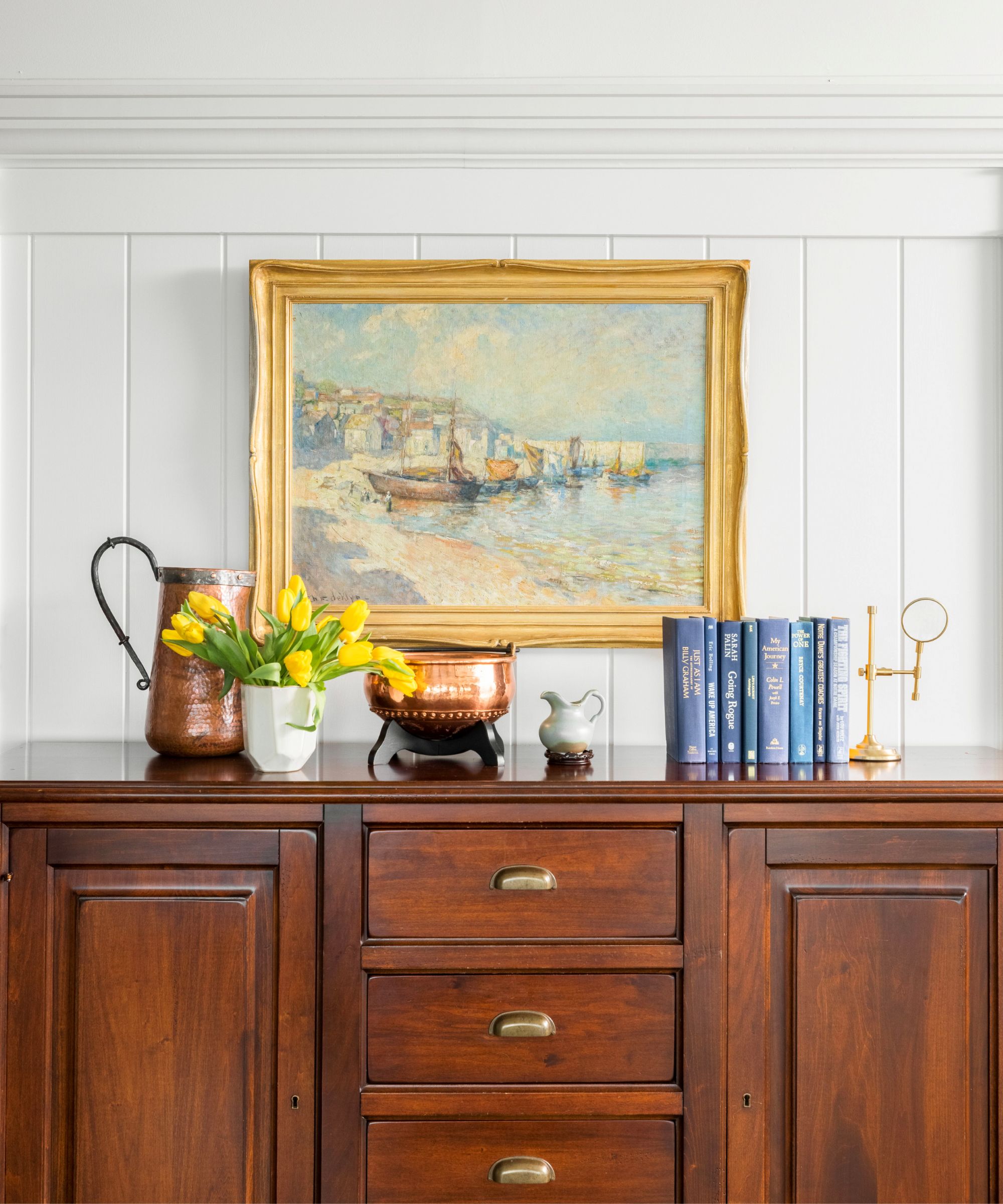 An antique dresser with flowers, books and trinkets on top, and a gold framed painting hanging above