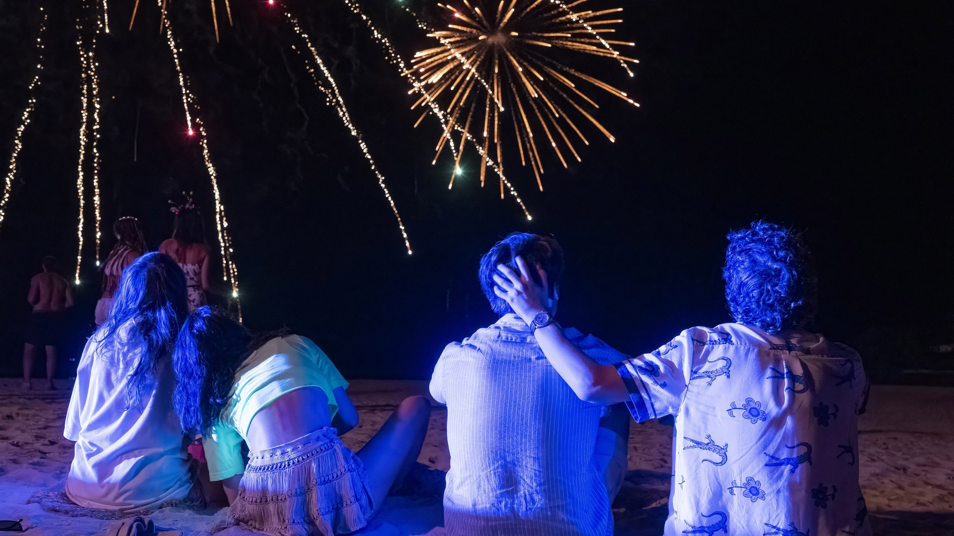 Aimee Lou Wood, Charlotte Le Bon, Patrick Schwarzenegger, Sam Nivola sit on a beach looking at fireworks