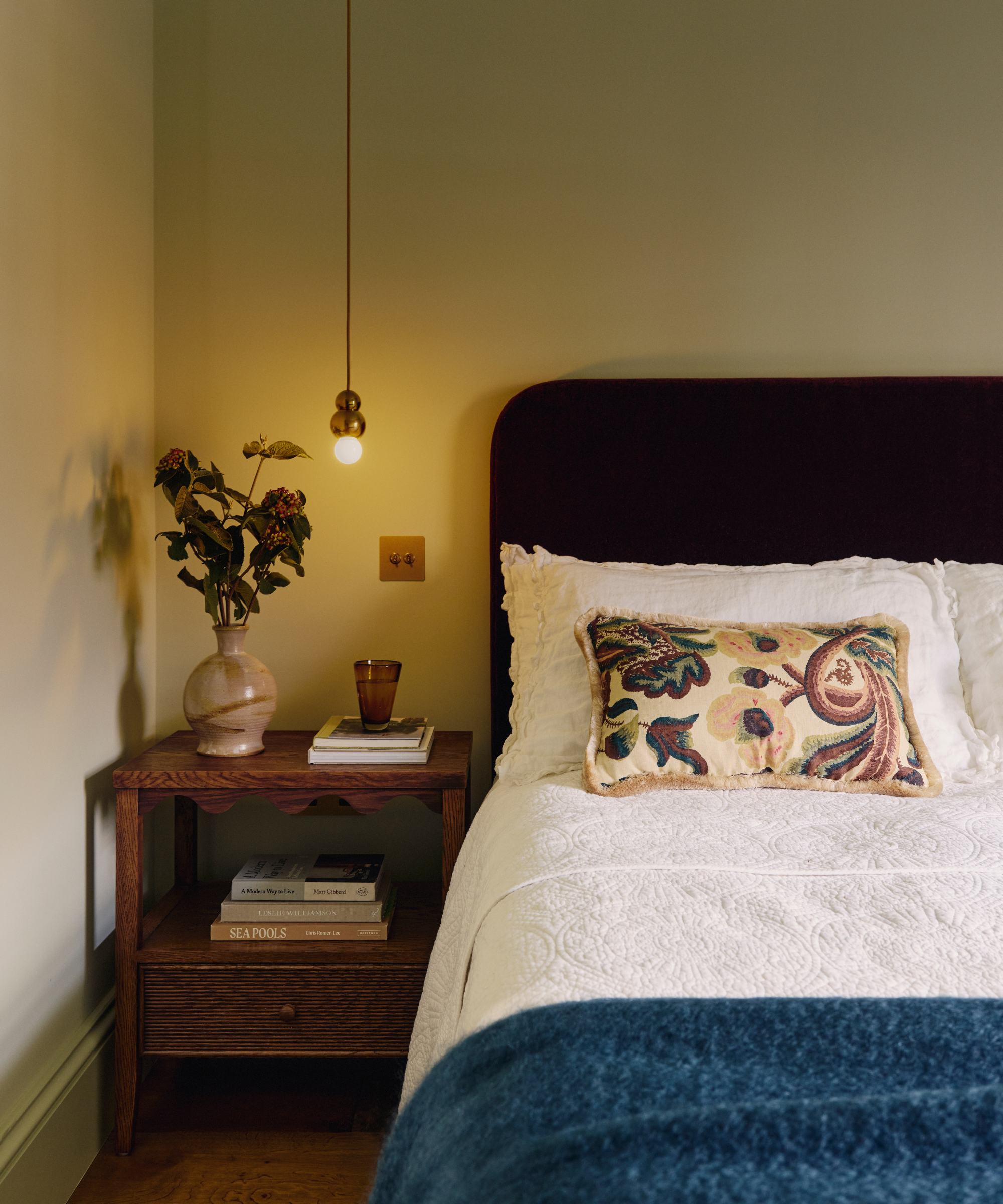 a bedroom with a small side table with decor on top, a blue blanket at the foot of the bed, and a decorative pillow