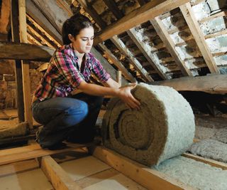 woman with dark hair in ponytail wearing check shirt and jeans rolling out loft insulation in loft space