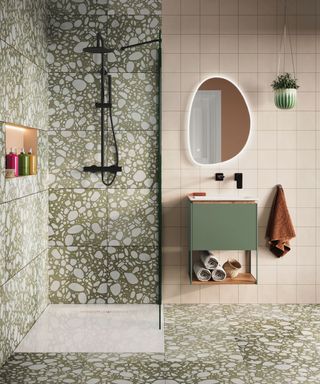 A bathroom with sage green and white dotted tiles, a shower, and a sage green sink unit with pastel pink tiles around it and a blob mirror above it