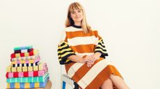 A young woman dressed in a stripy orange, cream, black, and yellow maxi dress sits next to a pile of towels in a similar pattern.