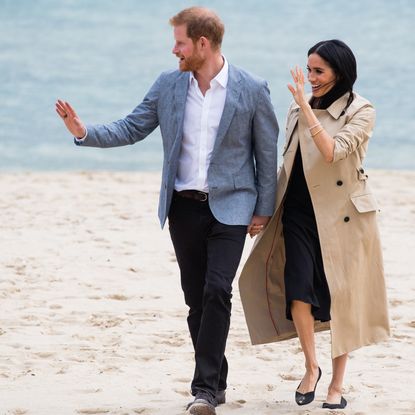 Meghan Markle wearing a tan trench coat and black dress and Prince Harry in black pants and a blue suit jacket walking on the beach and waving while holding hands