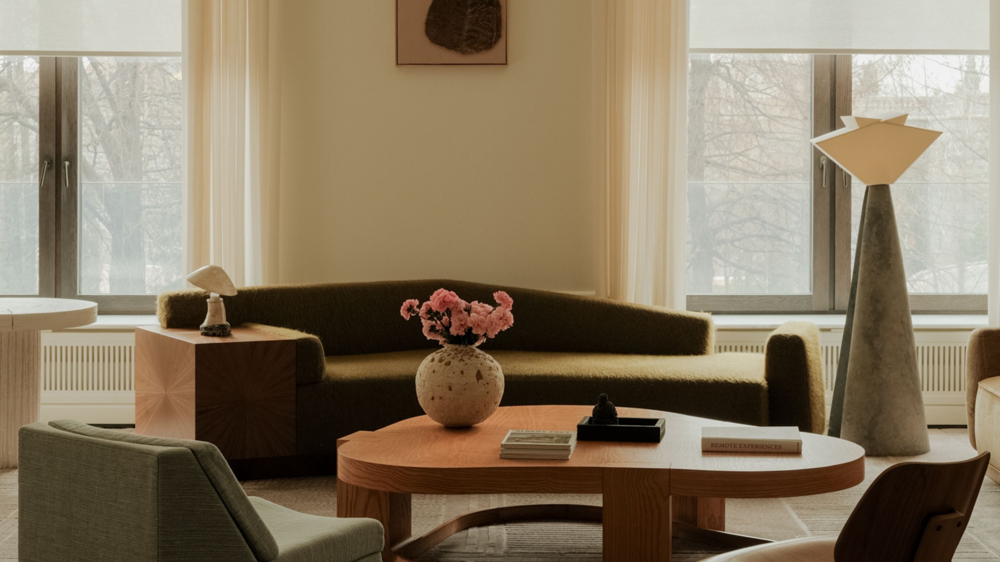 Image of a spacious living room that has a minimalist-meets-mid-century style. There are off-white walls with two large windows, a geometric, olive green sofa, an organic wooden coffee table, and a metal floor lamp.