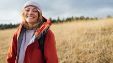 A happy woman in hiking clothes stands in front of an open field. She wears a beanie and is smiling widely.