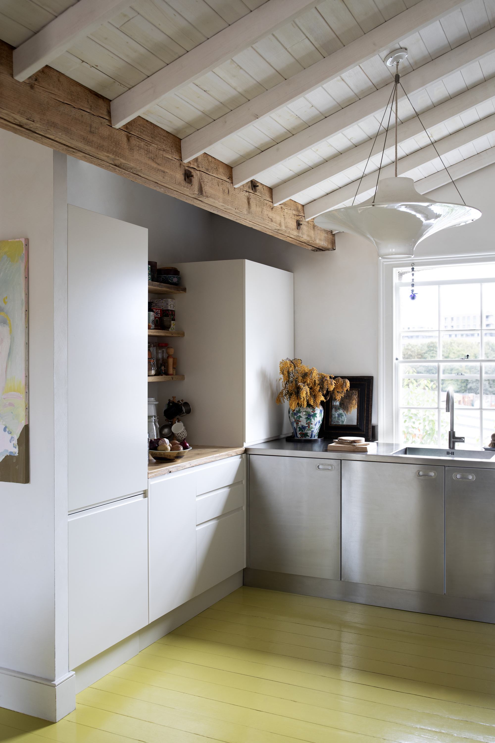 modern white kitchen with paneled ceiling, stainless cabinetry, timber shelves above nook, with yellow timber floorboards, pendant light, window over the sink, and pot of flowers