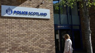 The Police Scotland logo and name on a sign fixed to a brick wall. A woman walks past it to the right, facing away from the camera.