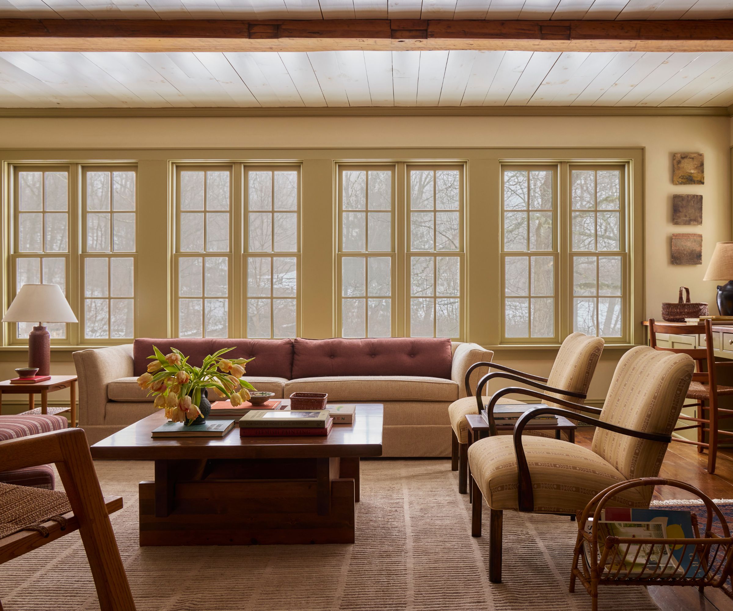 Warm living room with large butter yellow windows, a large beige sofa with pink cushions, two midcentury armchairs, a wooden coffee table, a striped rug, and a lamp in the corner.