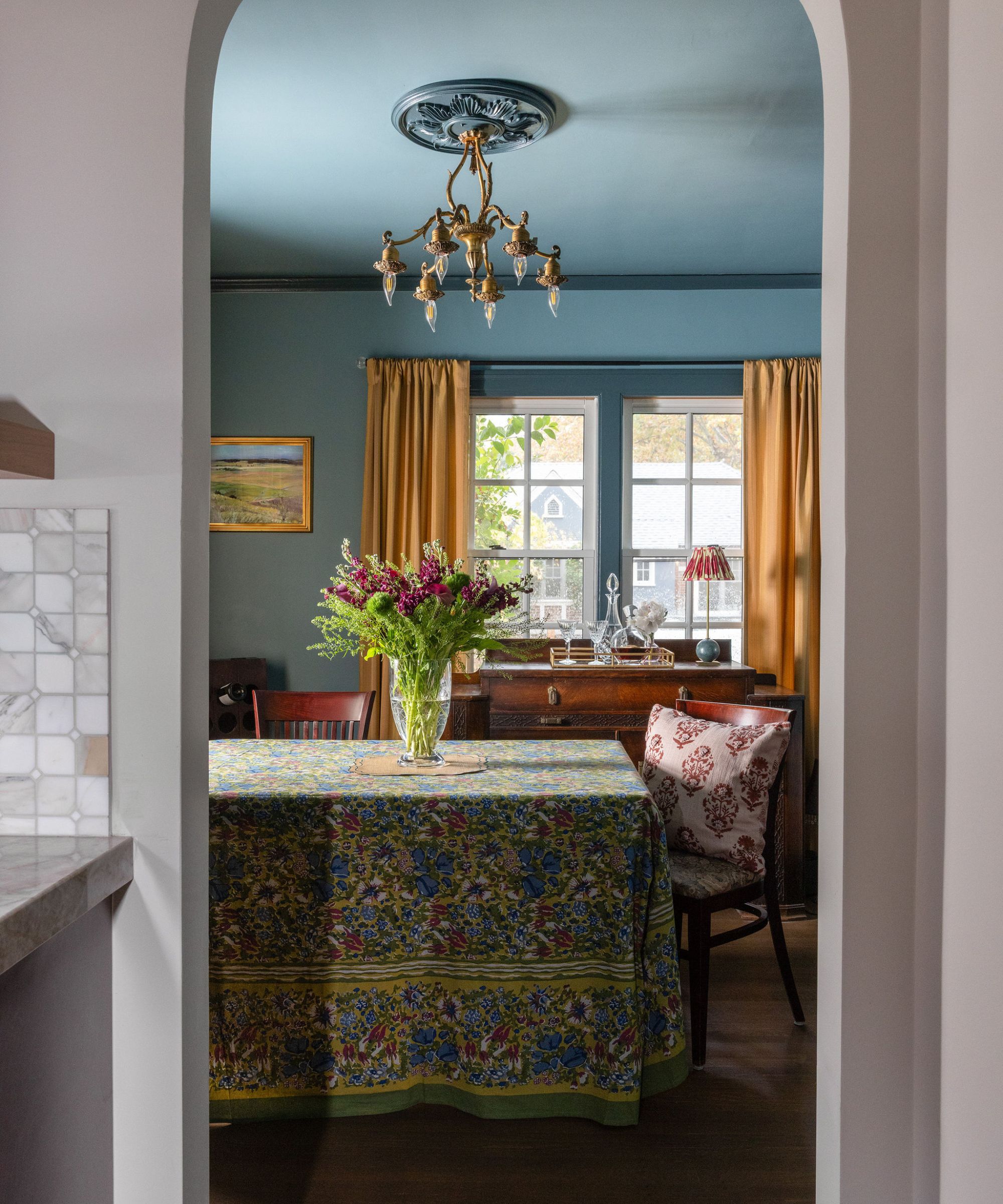 A dining room with teal walls, dark blue trim, mustard curtains, and a blue patterned tablecloth.
