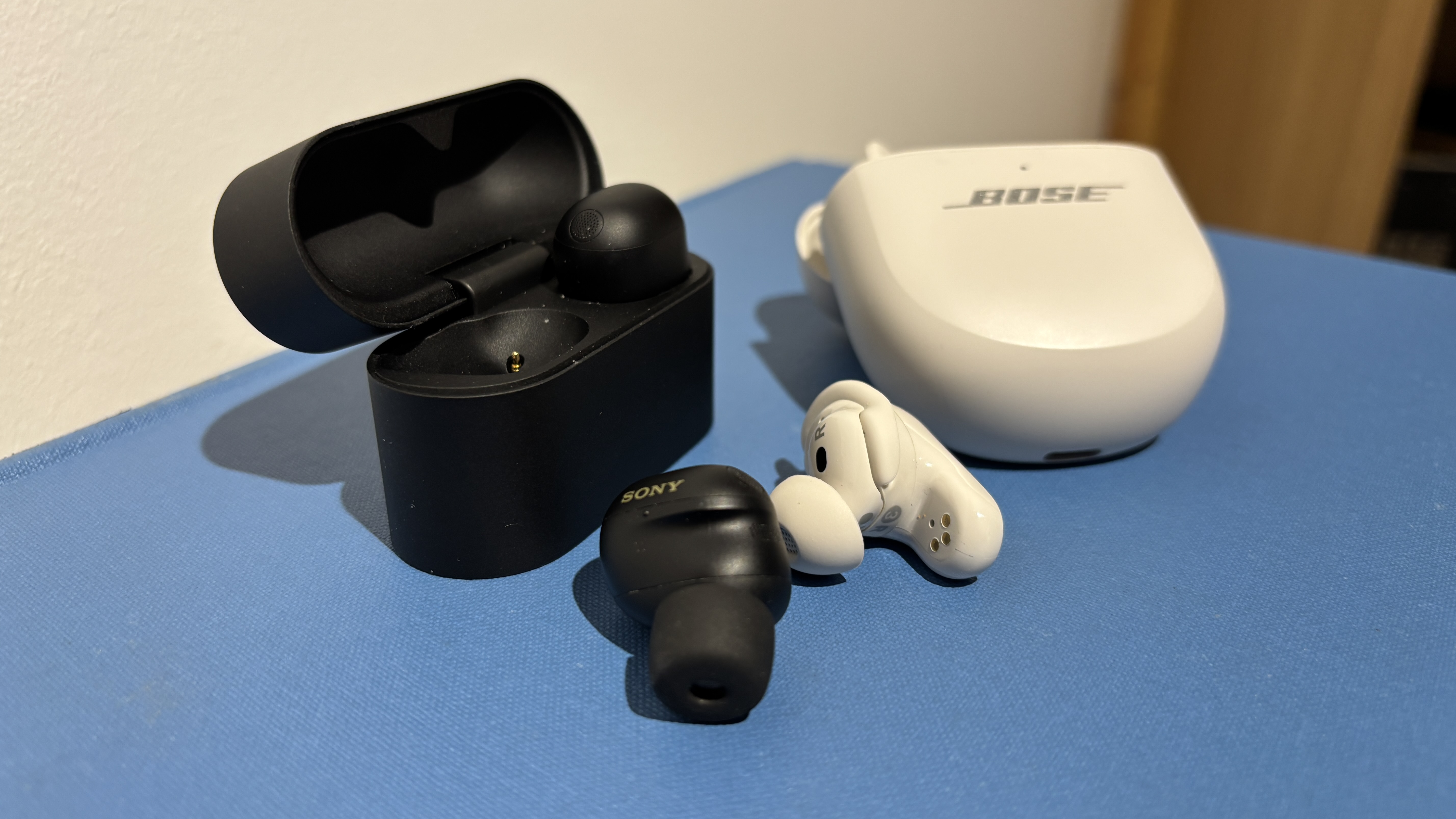 Sony WF-1000XM6 earbuds and Bose QC Ultra Earbuds (2nd Gen) on a blue surface with one earbud out of each case