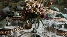 green themed table laid for thanksgiving with a white and green tablecloth, fall foliage centerpiece, wicker platemats and little velvet pumpkins