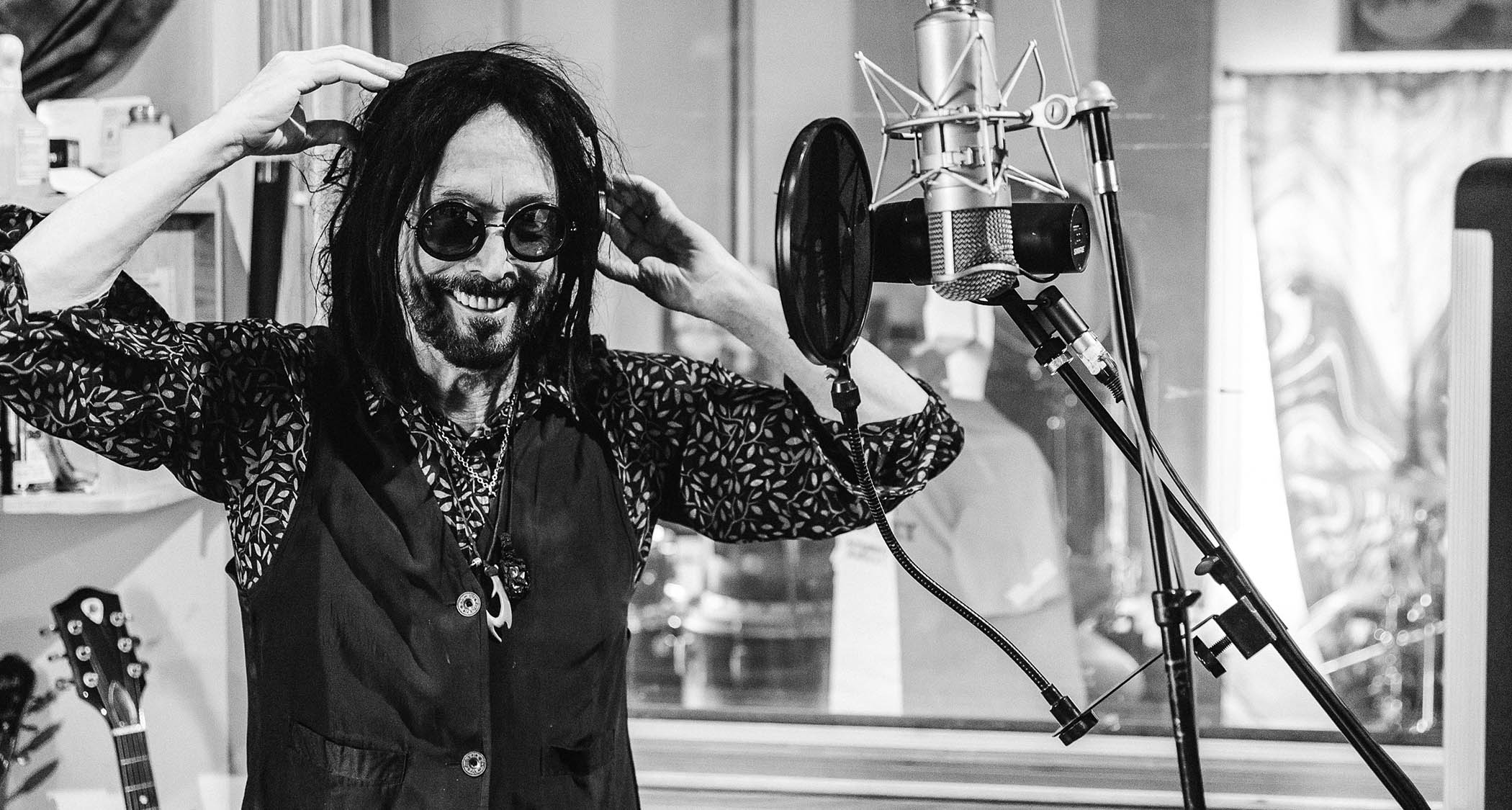 Mike Campbell has some fun in the vocal booth. This black and white image pictures him with a patterned shirt, black waistcoat and dark sunglasses.
