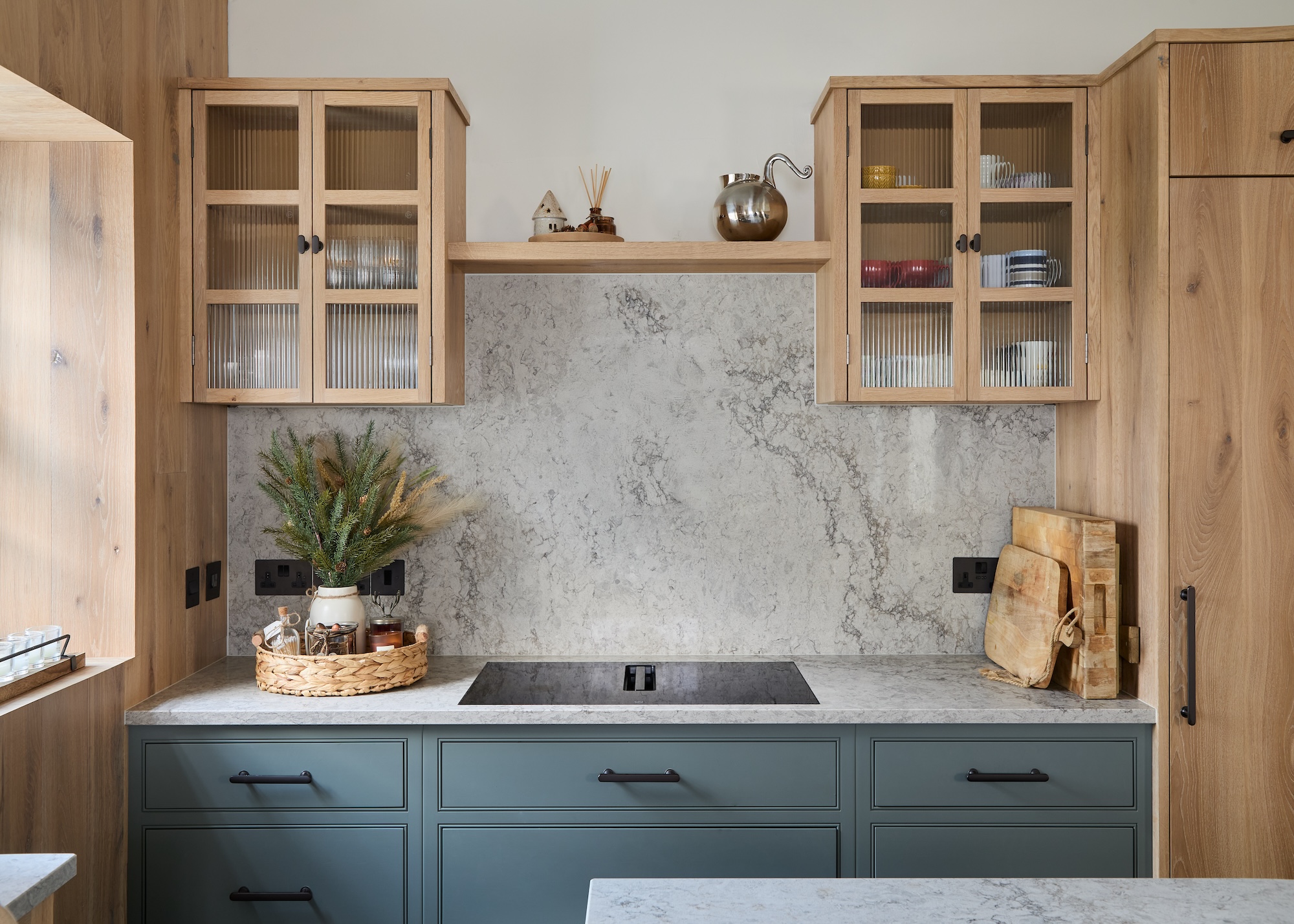 An induction hob on top of a kitchen counter and above the counters are glass fronted cabinets on either side of it. Beside the hob is a tray with flowers as well as kitchen essentials and on the other side are two chopping boards