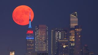 The full Buck Moon or Thunder Moon (a red full moon) passes behind Hudson Yards and the Empire State Building lit in the flag colors of countries competing in the Tokyo Olympics as it rises in New York City on July 23, 2021 as seen from Lyndhurst, New Jersey.