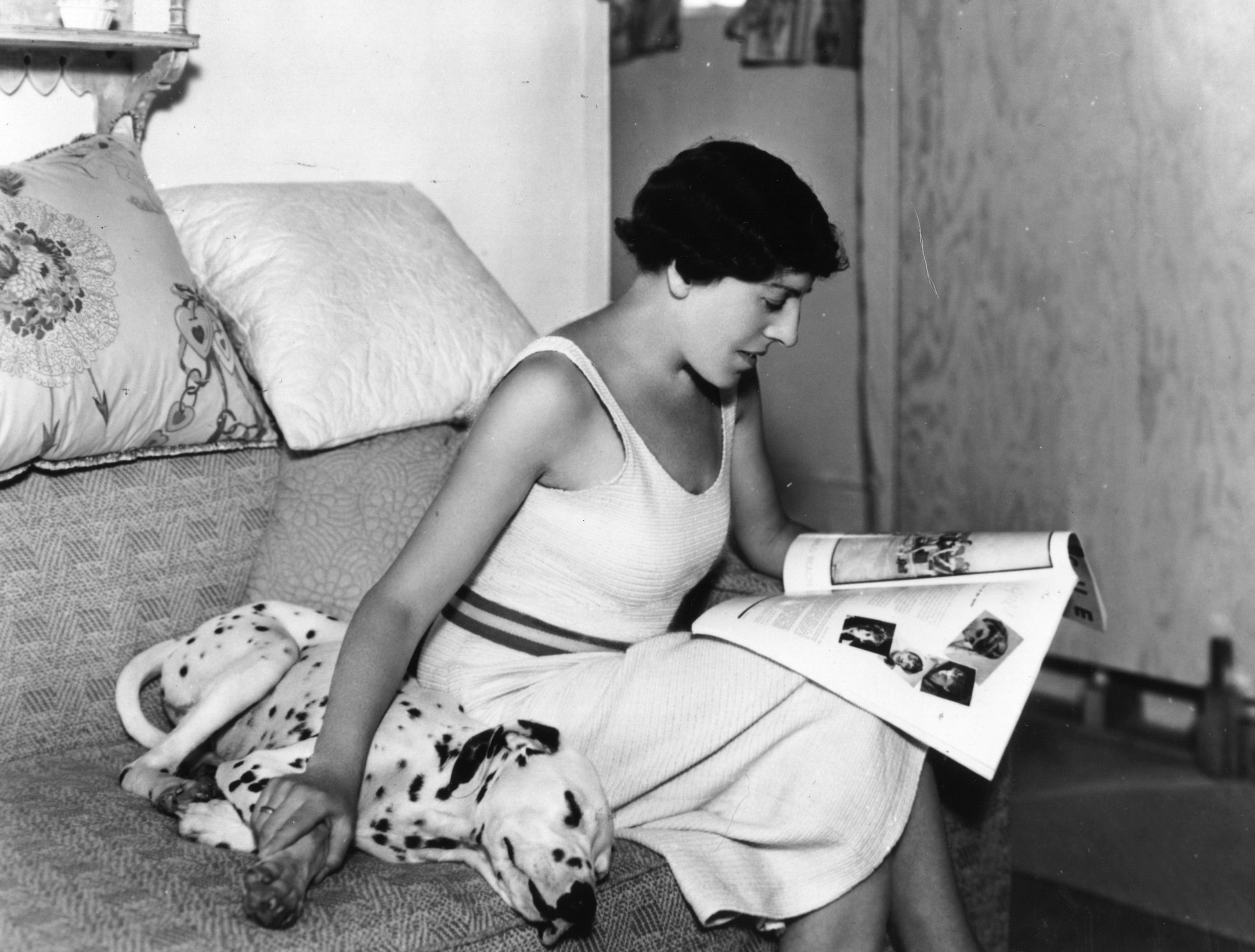 Author Dodie Smith reading a magazine with one hand and stroking a sleeping Dalmatian dog with the other