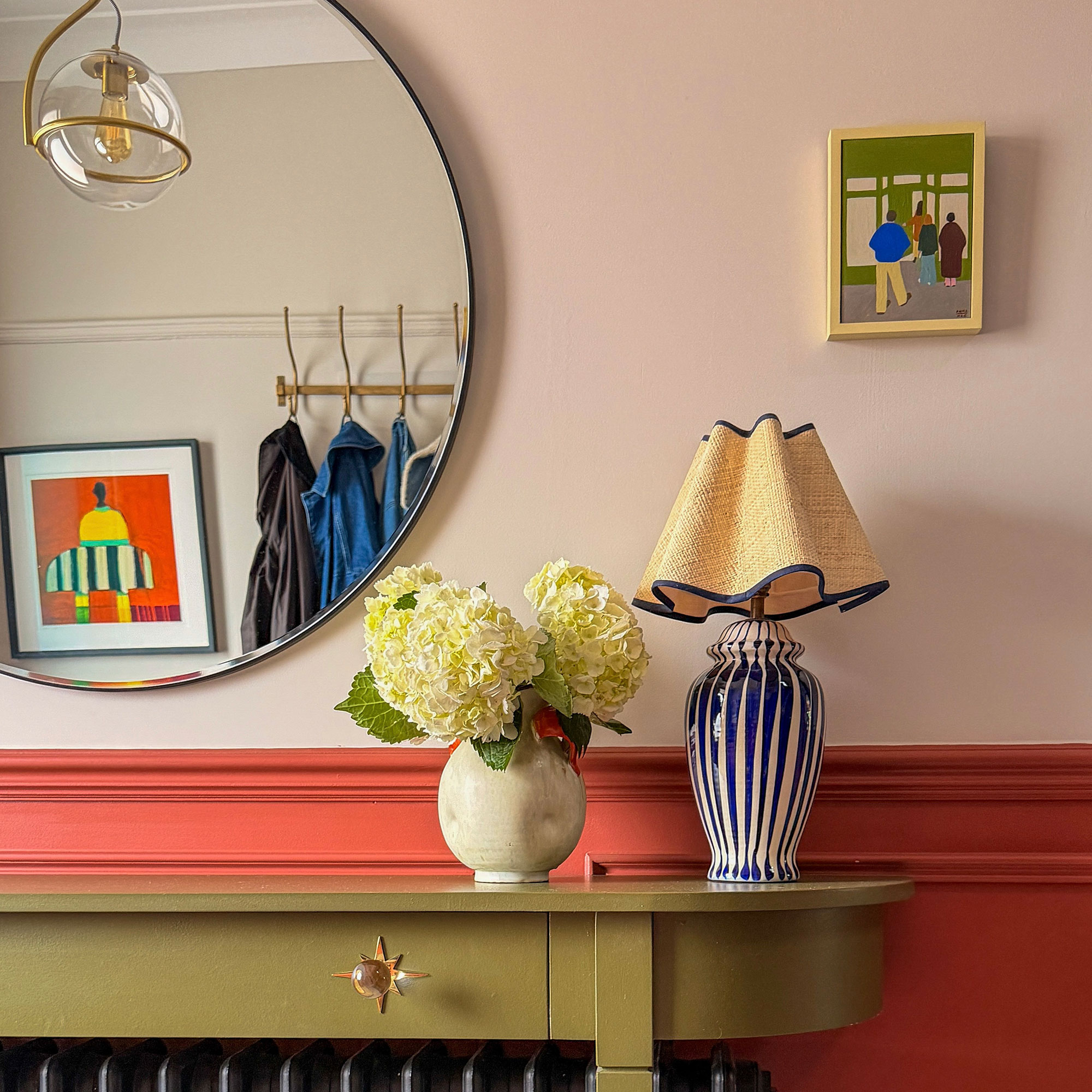 Hallway with lower half painted coral and top salmon, with green radiator cover console table with vase of hydrangeas and blue and white striped lamp, beneath round mirror reflecting coat rack