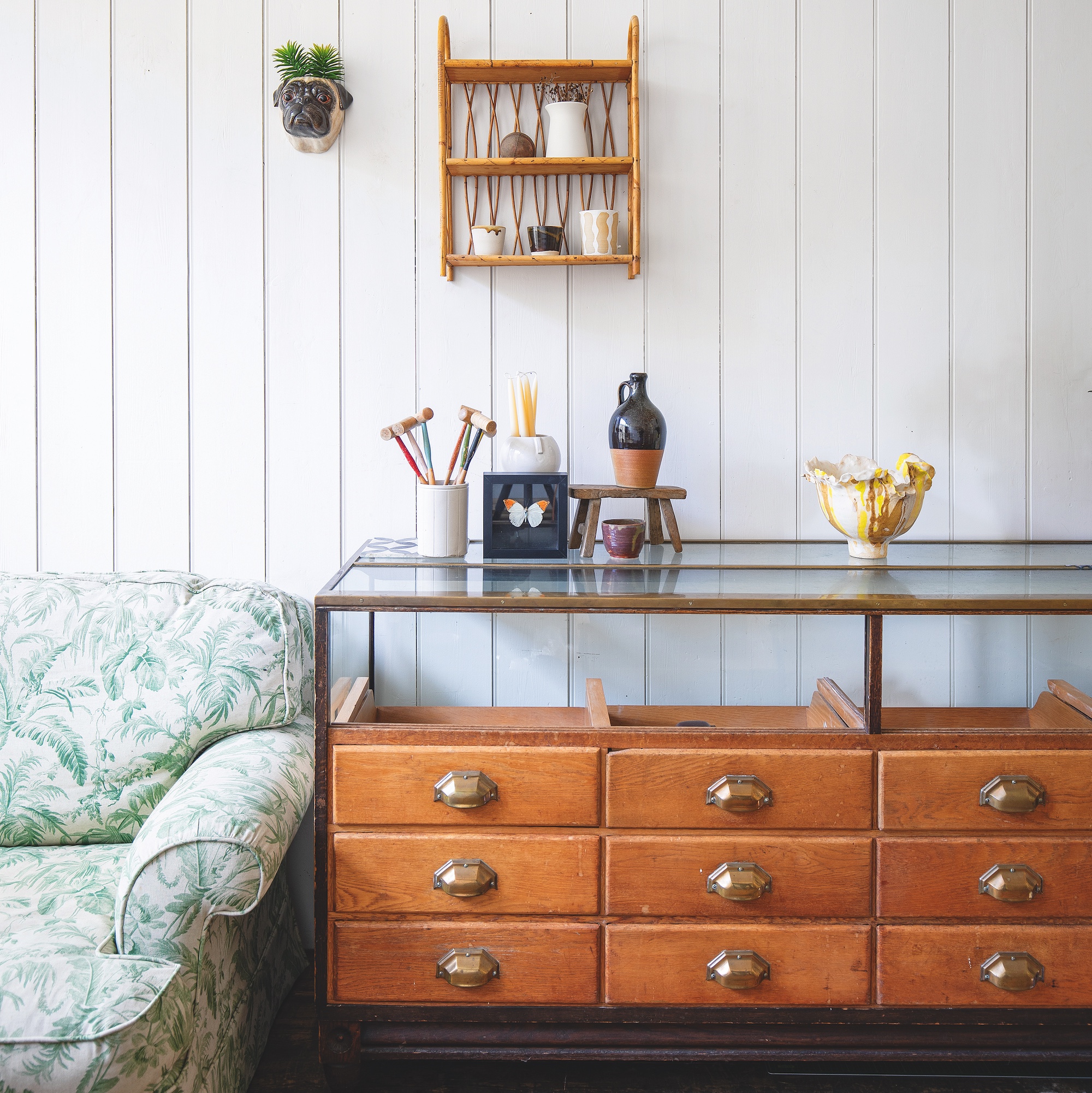 room with white painted wood panel walls, green floral sofa and large haberdashery cabinet