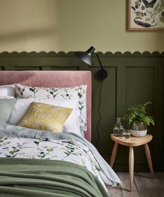 Bedroom with dark green wall paneling with scalloped edging, light green painted walls, black wall lamp, light wooden stool, green and grey floral bedding with pale pink headboard, scatter cushions