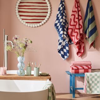 A white round bobbin mirror hangs on a pink wall next to patterned towels