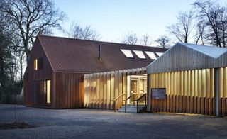 Daylight image, wooden barn type building, with skylight roof and thin chimney, adjacent building attached with wooden slat front and roof, lights glowing through the openings, small set of stone steps leading up to the entrance door, with handrails, building information sign to the right, gravel driveway, winter trees in the distance, clear pale blue sky