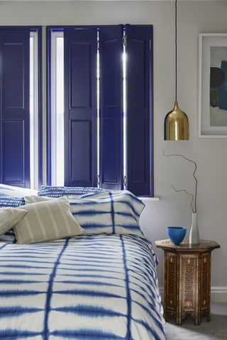 bedroom with blue shutters, blue and white tie-dyed duvet, copper Moroccan side table, brass pendant light, artwork