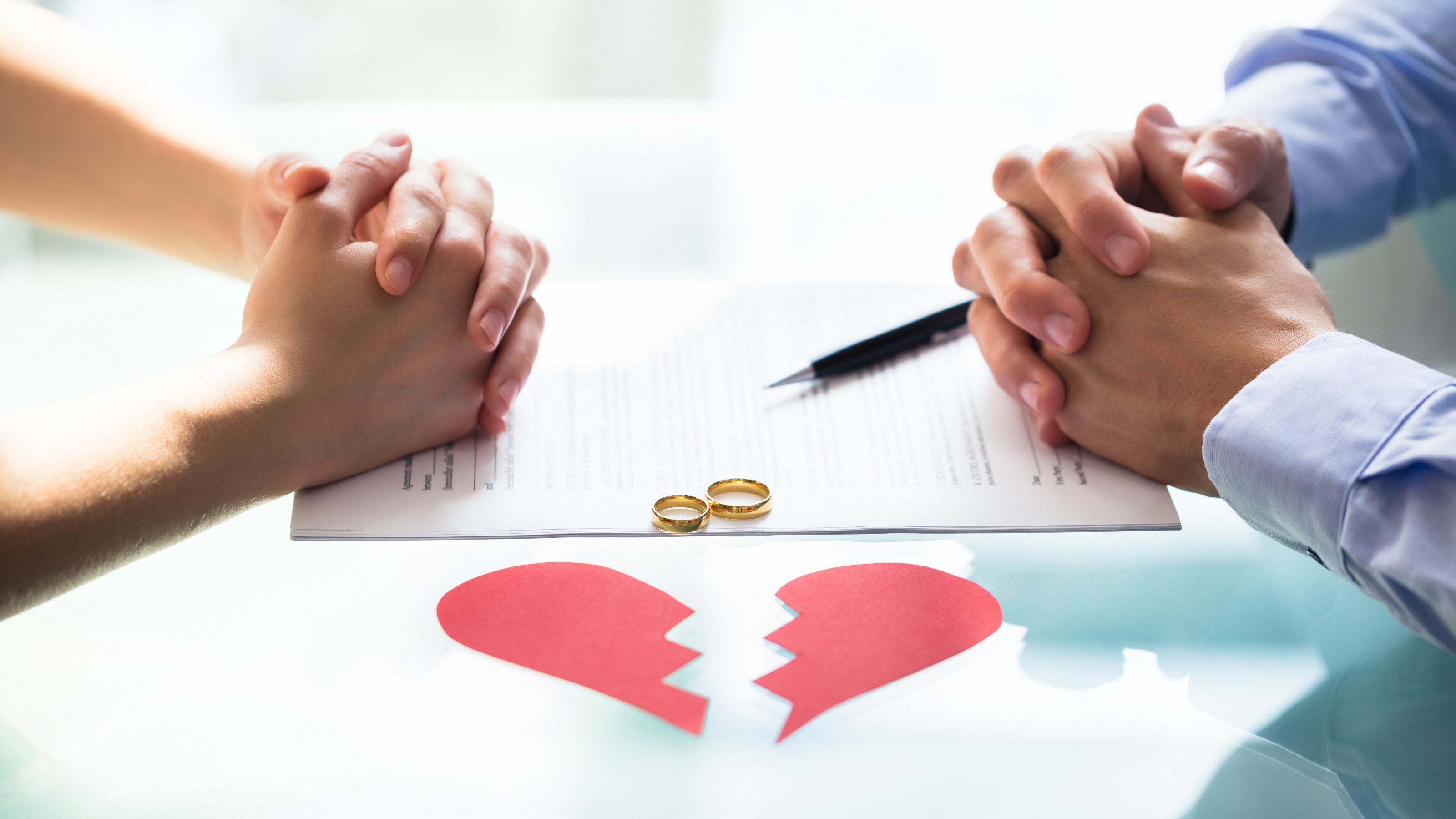 A divorcing man and woman sit across from each other with folded hands on top of paperwork, a broken heart made of construction paper between them.