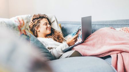 A woman with long, curly hair is shown relaxing on a gray sofa, partially covered by a pink blanket. She is leaning back against pillows while holding a smartphone in one hand and resting a laptop on her lap. She has one hand behind her head and a soft smile on her face, suggesting a comfortable, informal work-from-home or leisure environment.