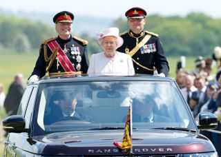 Queen Elizabeth and two uniformed men riding in an open topped Range Rover wearing a pink coat