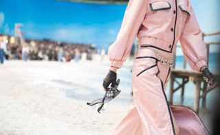 Model wears leather baby pink jacket and trousers and carries heeled sandals