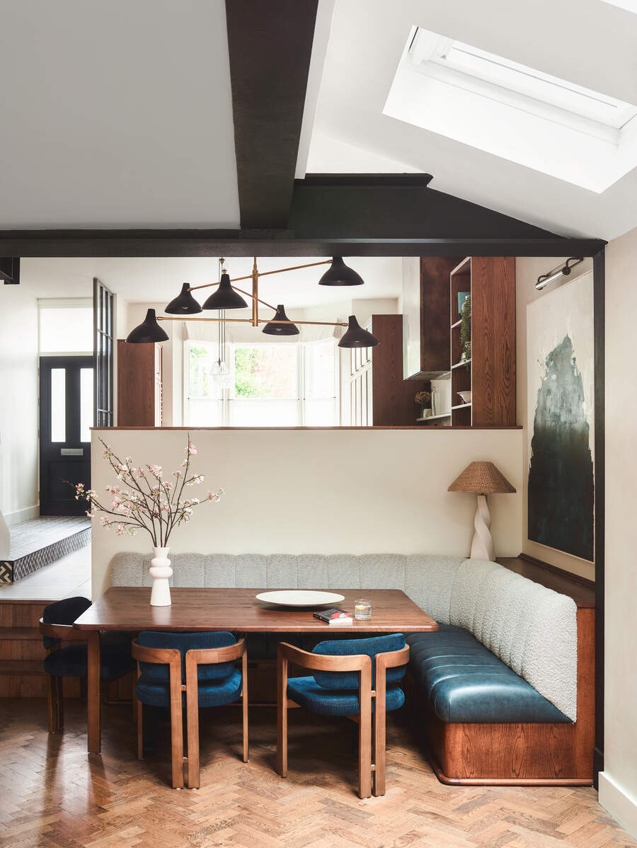Image of a dining nook that has open stairs up to a kitchen. The walls are beige and the ceiling is white with the trim painted black. The nook has a L-shaped booth seat that has a leather teal seat and a light blue boucle back.