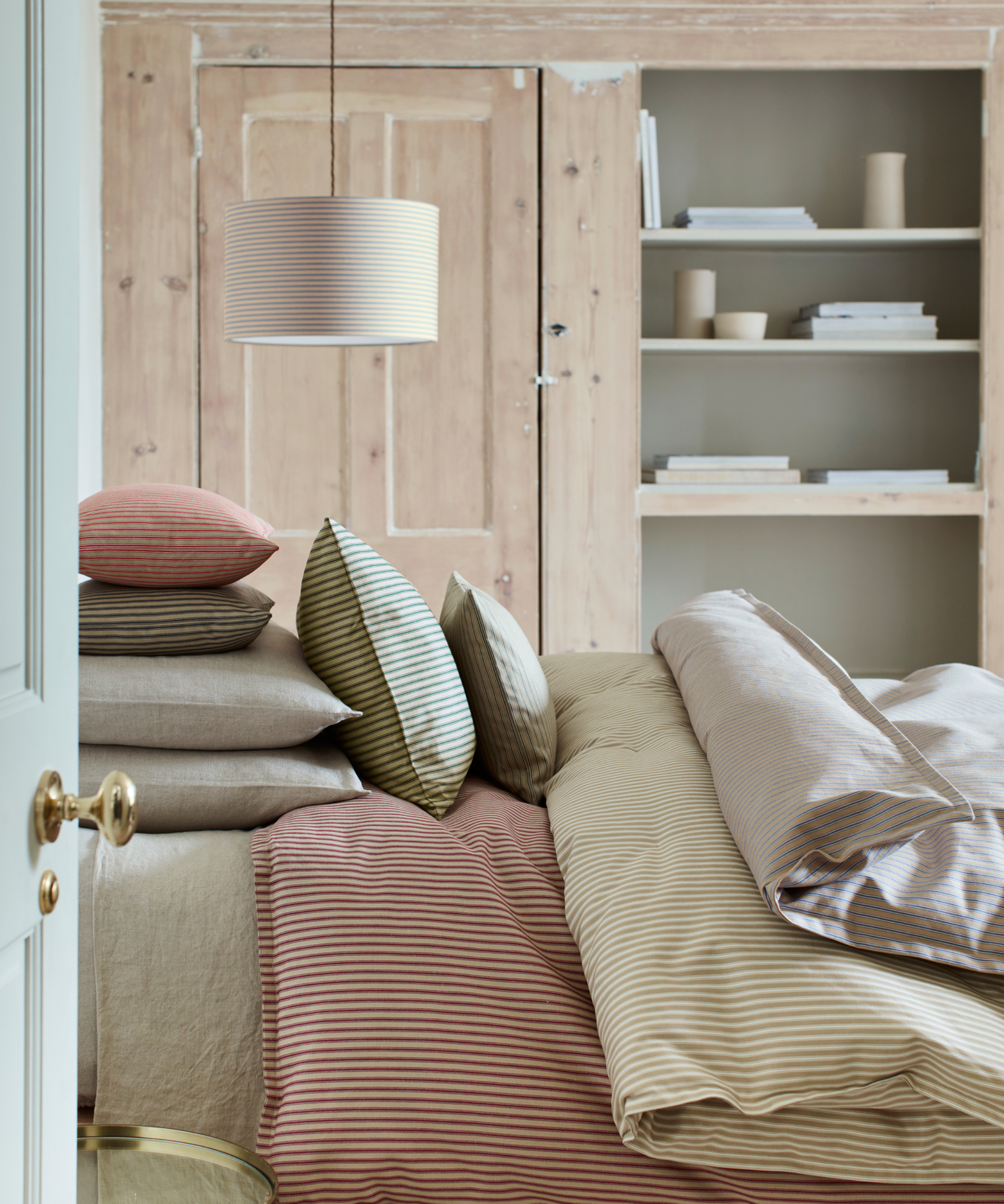 bedroom with striped bedlinen on bed and low hanging striped pendant light to one side of bed, door and recessed shelving on wall behind