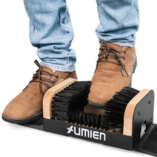 Bottom half of a person's legs in blue jeans and brown suede boots with one foot in the scraper that has two bristle brushes facing each other and space to put the boot in between