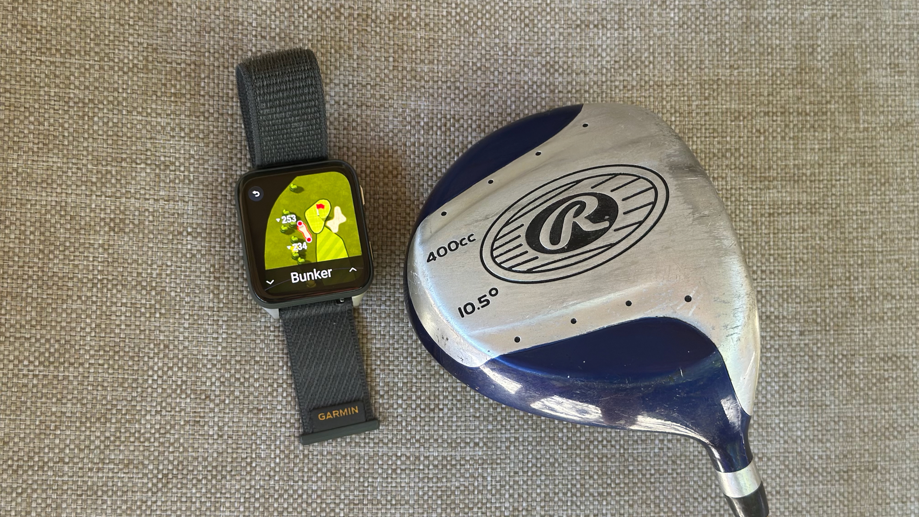 The Garmin Venu X1 showing a Golf activity with a highlighted bunker hazard and the distance to it; next to the watch is a driver head.