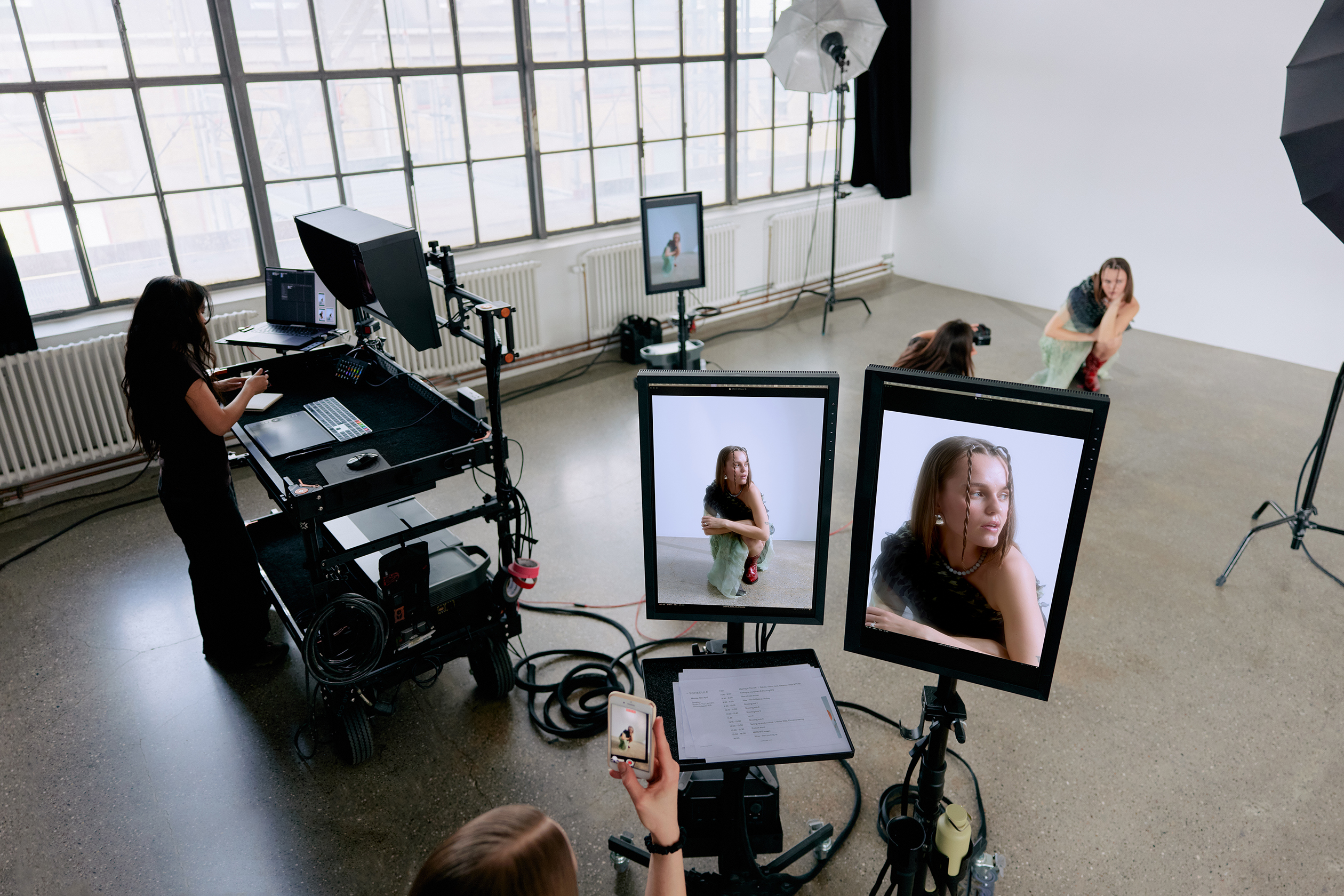 Photo studio with a model posing, two monitors showing her close-up, a photographer shooting, and a technician working at a control station