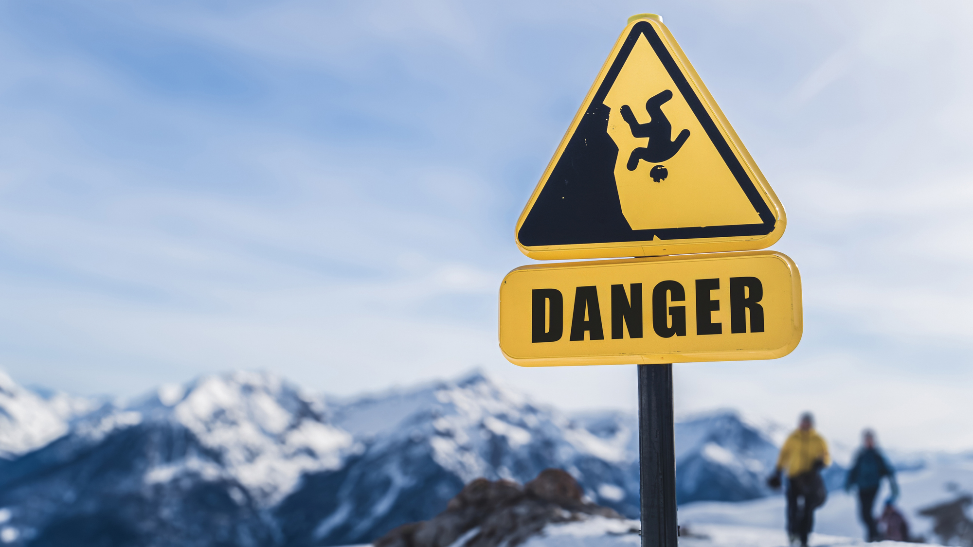 A yellow danger sign warns of steep cliffs in a snowy mountain landscape. Two people in winter gear are visible in the background, highlighting the hazardous conditions of the area