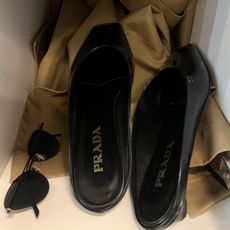 A pair of black Prada flat mules in a locker with sunglasses and a trench coat.