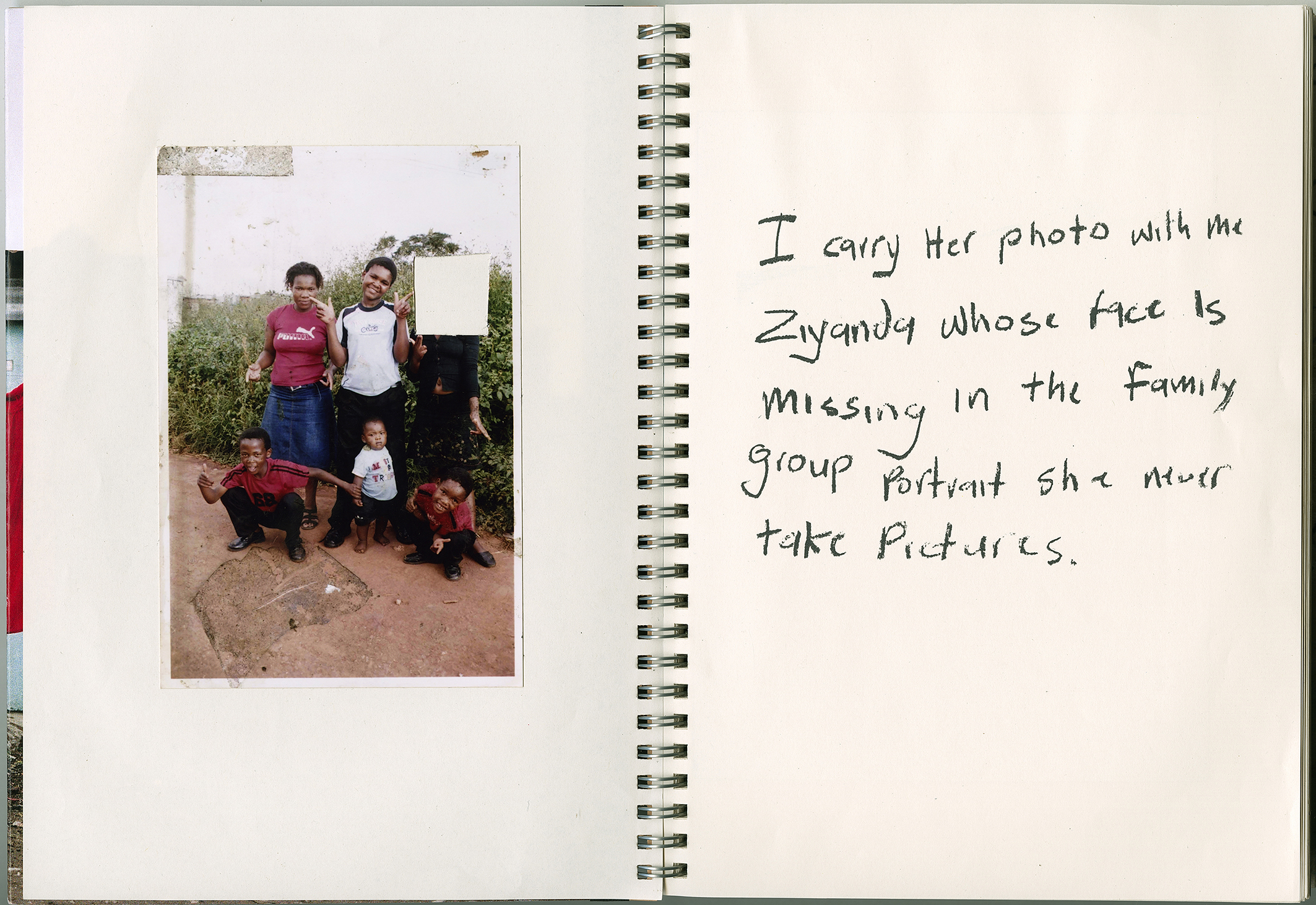 Color photograph of a notebook with an image of two women and three children crouched in front on the left hand side. On the right, the words "I carry her photo with me Ziyanda whose face is missing in the family group portrait she never take pictures." are written
