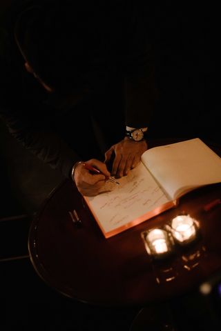Image of Marc Newson writing with a red fountain pen, in an open white page book, dimly lit room, round dark wood table, two glass tea light candle holders with lit candles, black backdrop