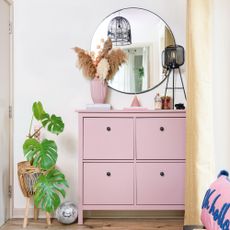 White hallway with pink shoe storage, a round mirror and dried flowers