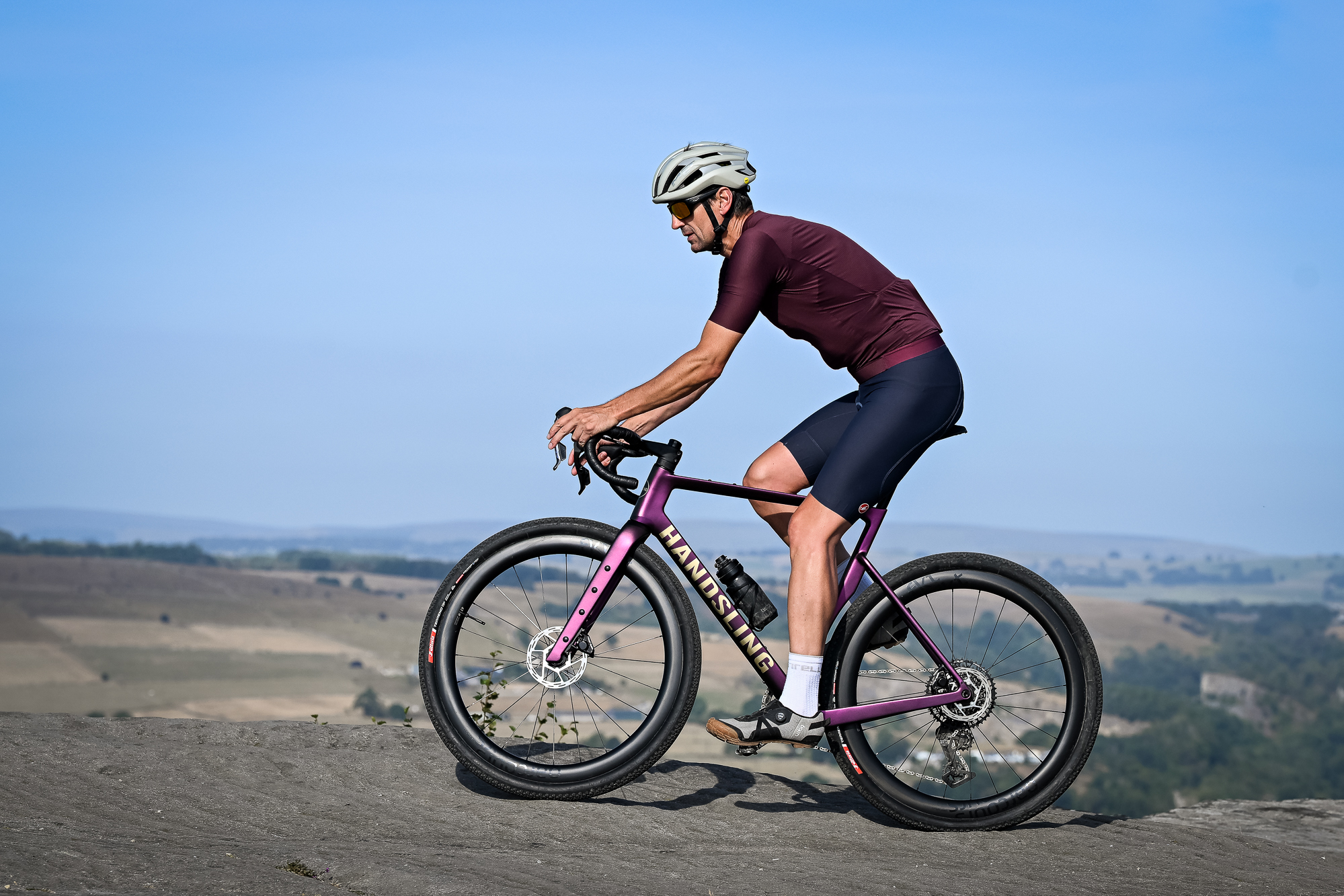 Man riding a purple gravel bike along a rocky edge wearing grey shoes, blue shorts, a purple top and a beige helmet.