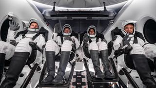 four astronauts and white and black spacesuits wave while sitting inside a space capsule.