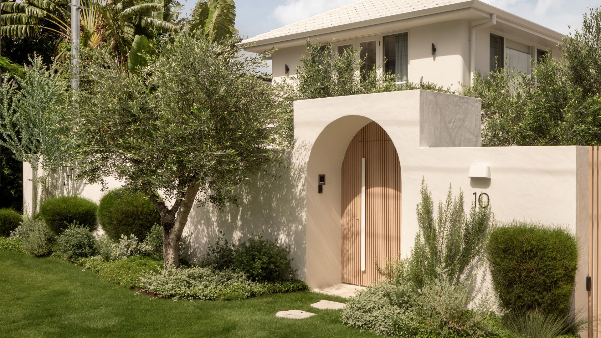 A modern home with a front yard with round topiaries, ground cover, a lawn, and olive trees by an arched doorways with a wood panelled door