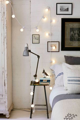 White bedroom with exposed brick wall and fairy lights, gallery wall, black stool with gray angle poise lamp, books